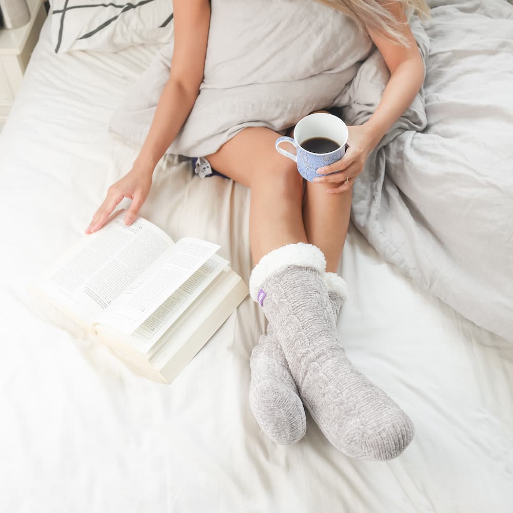The Foozie™ in Light Gray, worn in bed with a book and a cup of coffee