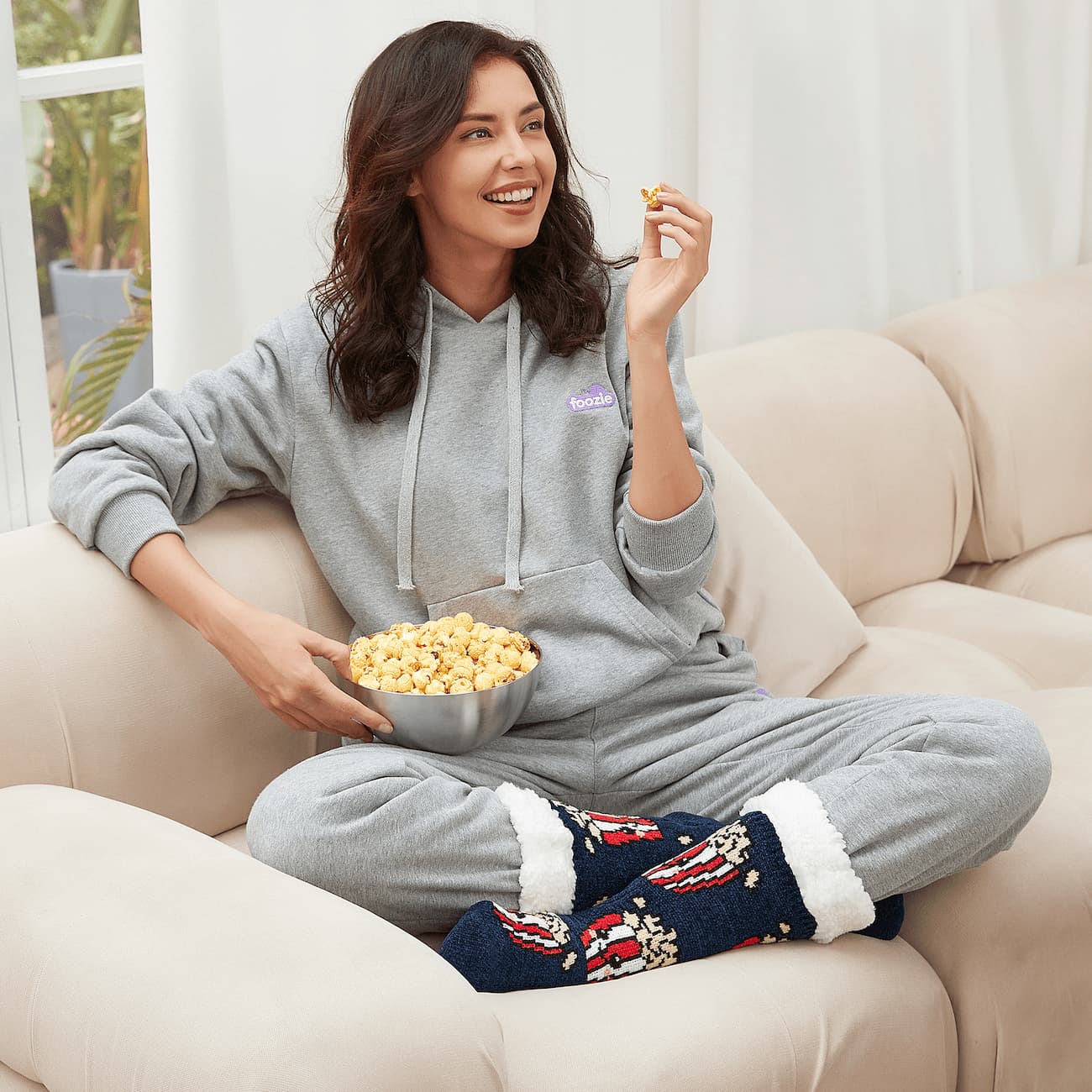 The Foozie™ Popcorn slipper socks, worn by a woman in a hoodie on the sofa with a bowl of popcorn